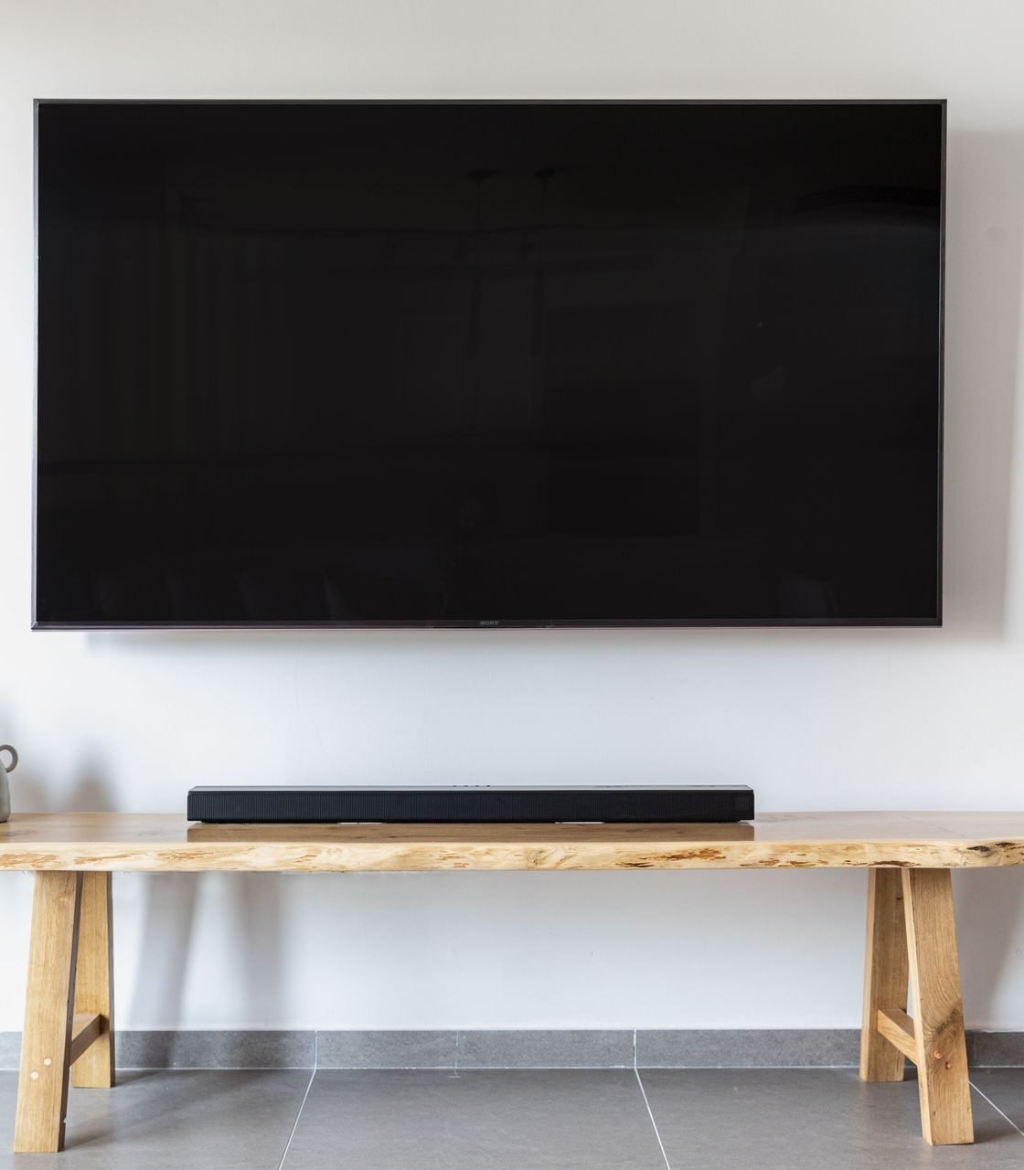 Soundbar sobre banco de madeira abaixo de uma TV fixada na parede branca