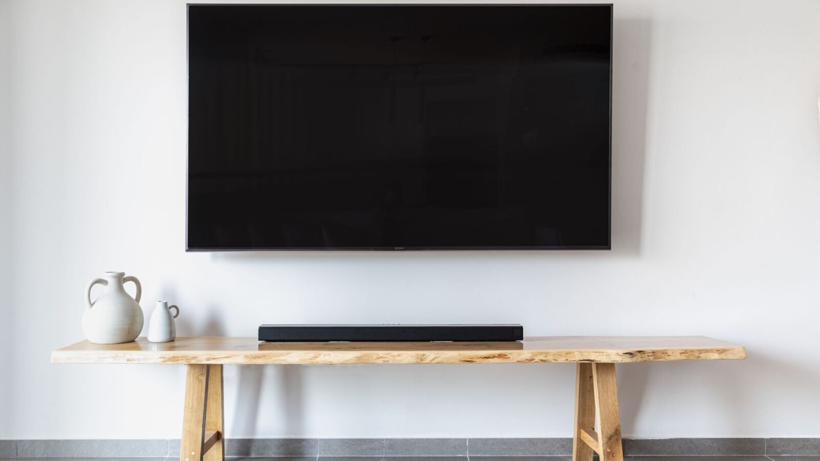 Soundbar sobre banco de madeira abaixo de uma TV fixada na parede branca