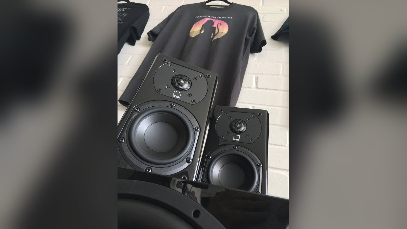 História da SVS: caixas de som SVS pretas, com logo visível, em frente a camisetas penduradas em parede de tijolos brancos