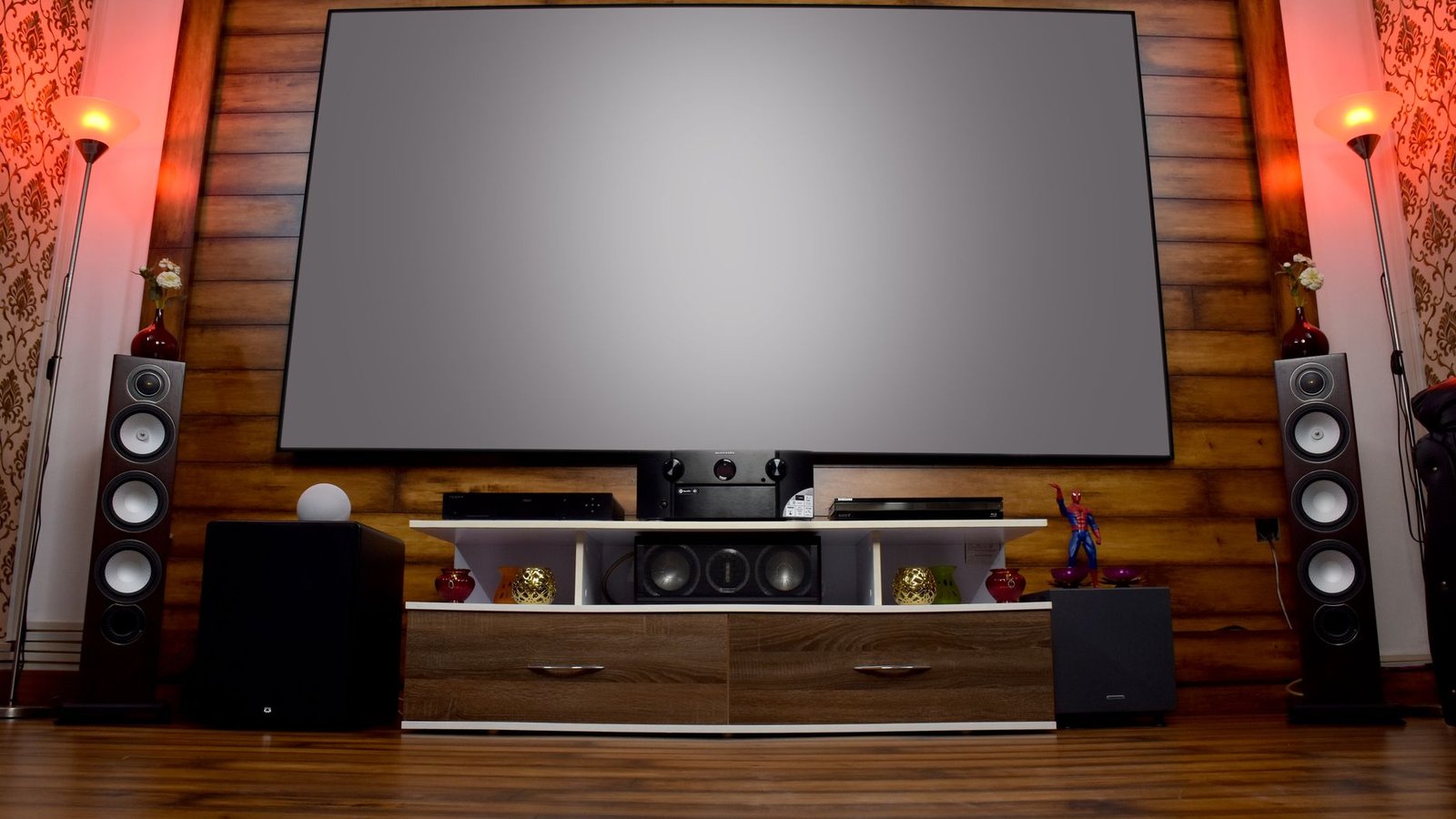 Sala de estar com TV de tela grande montada em parede de madeira, soundbar preta central sobre rack, duas caixas acústicas torre altas laterais, subwoofers e iluminação ambiente vermelha