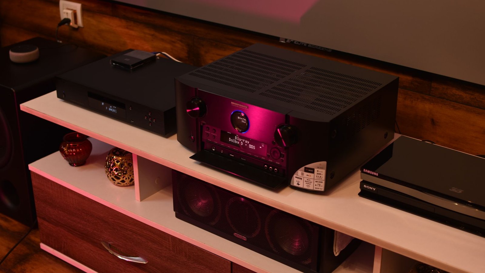 Receptor AV Marantz com iluminação roxa, posicionado em estante de home theater ao lado de outros componentes como Blu-ray player e caixas acústicas