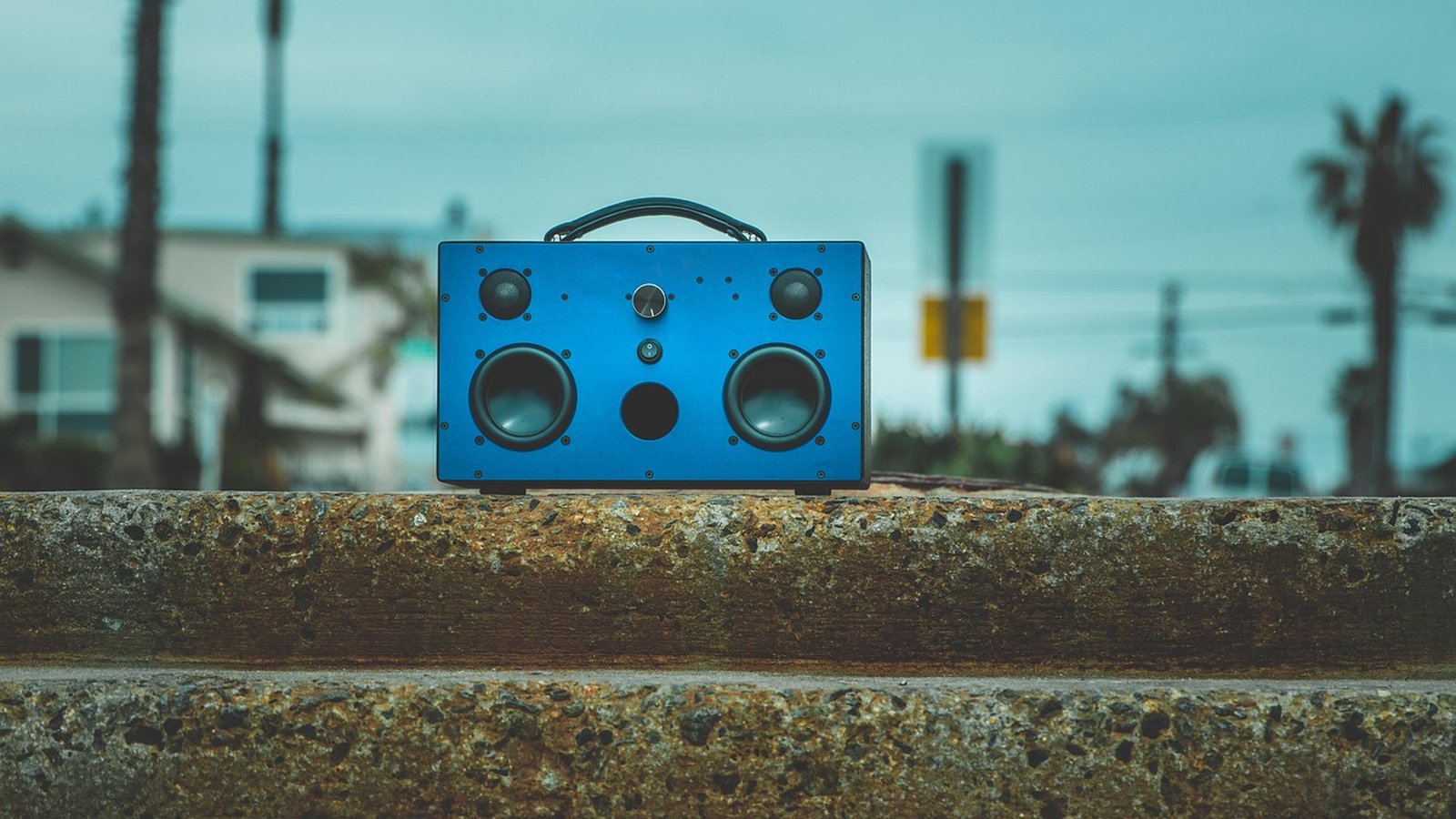 Caixa de som portátil azul robusta, com dois alto-falantes principais, dois tweeters e controles na frente, posicionada sobre uma parede de concreto em ambiente externo