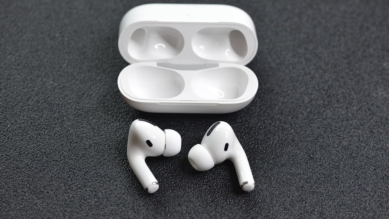 Fones de ouvido intra-auriculares sem fio (TWS) brancos, modelo AirPods Pro, com estojo de carregamento aberto, sobre superfície escura texturizada. O foco está nos fones e no estojo, destacando o des