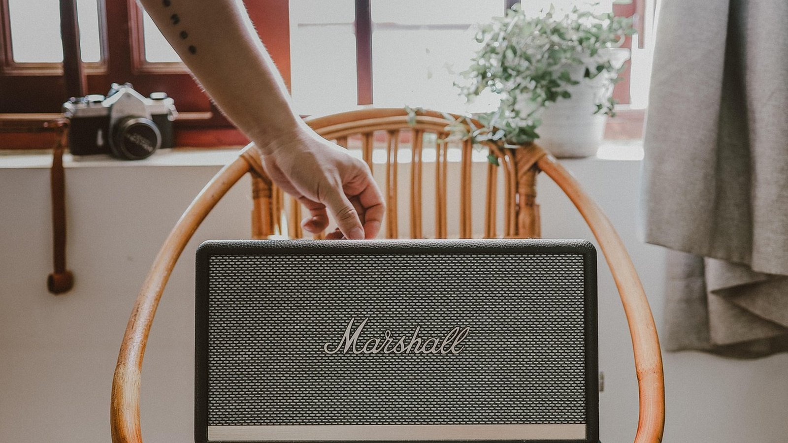 Caixa de som Marshall preta com grade texturizada, posicionada sobre uma cadeira de madeira rústica, com mão ajustando o aparelho