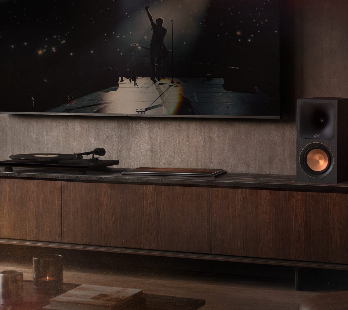 Par de caixas ativas Klipsch RP-500PM, com woofers cor de cobre, sobre um rack de madeira ao lado de um toca-discos, abaixo de uma TV