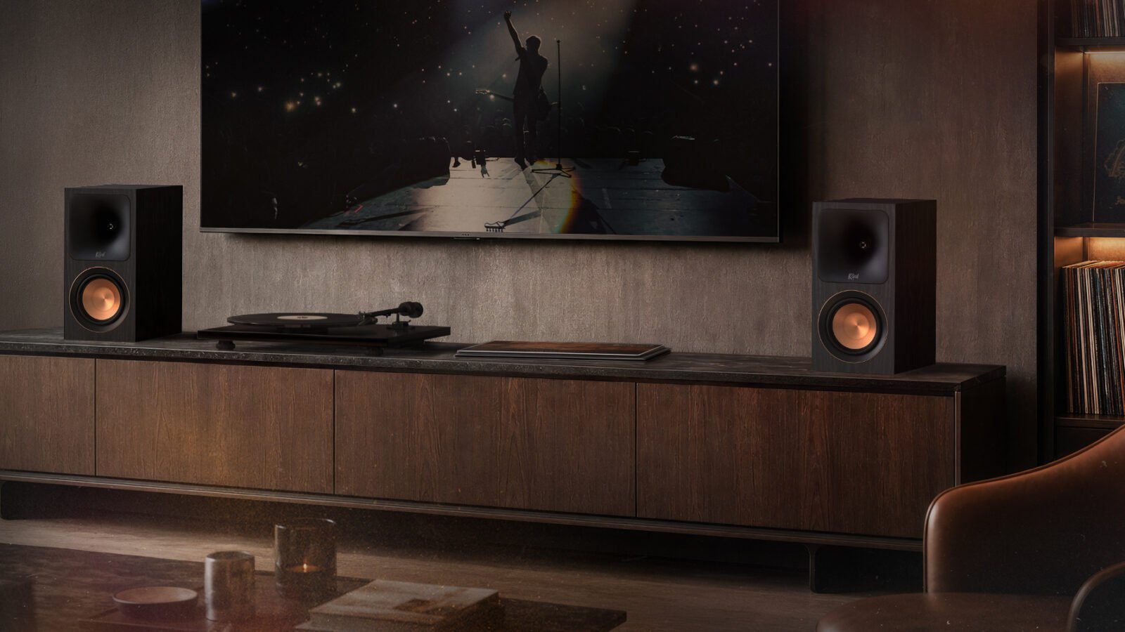 Par de caixas ativas Klipsch RP-500PM, com woofers cor de cobre, sobre um rack de madeira ao lado de um toca-discos, abaixo de uma TV