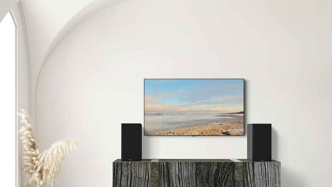 Par de caixas bookshelf Klipsch RP-600M II sobre um móvel de madeira escura, ao lado de uma TV