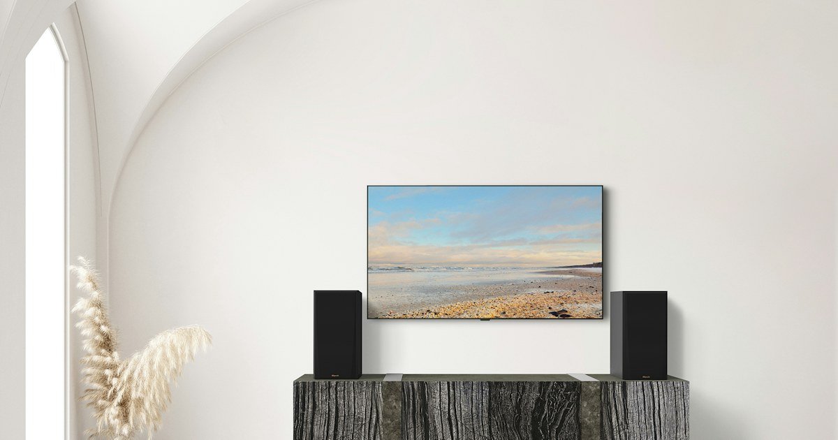 Par de caixas bookshelf Klipsch RP-600M II sobre um móvel de madeira escura, ao lado de uma TV