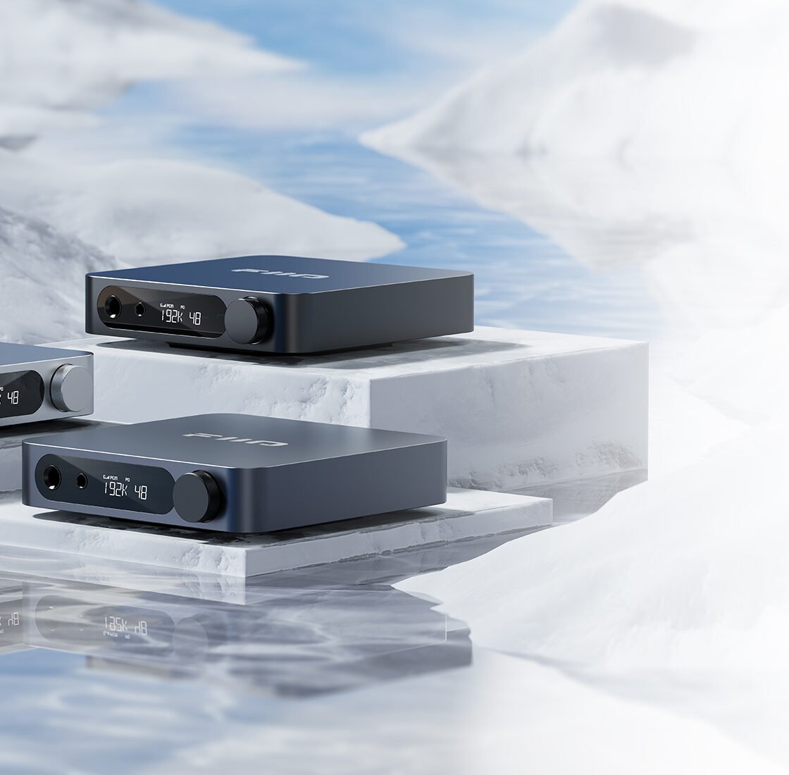 Três unidades do DAC/amplificador de mesa FiiO K11 nas cores preta, prata e titânio sobre blocos brancos