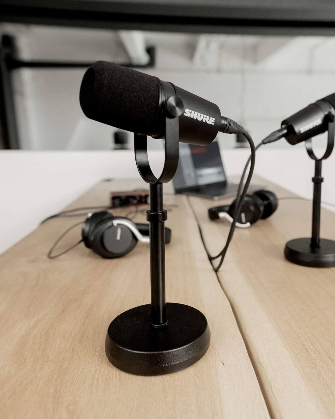 Dois microfones Shure MV7X pretos em pedestais de mesa, conectados por cabo XLR, sobre bancada de madeira com fones e interface de áudio