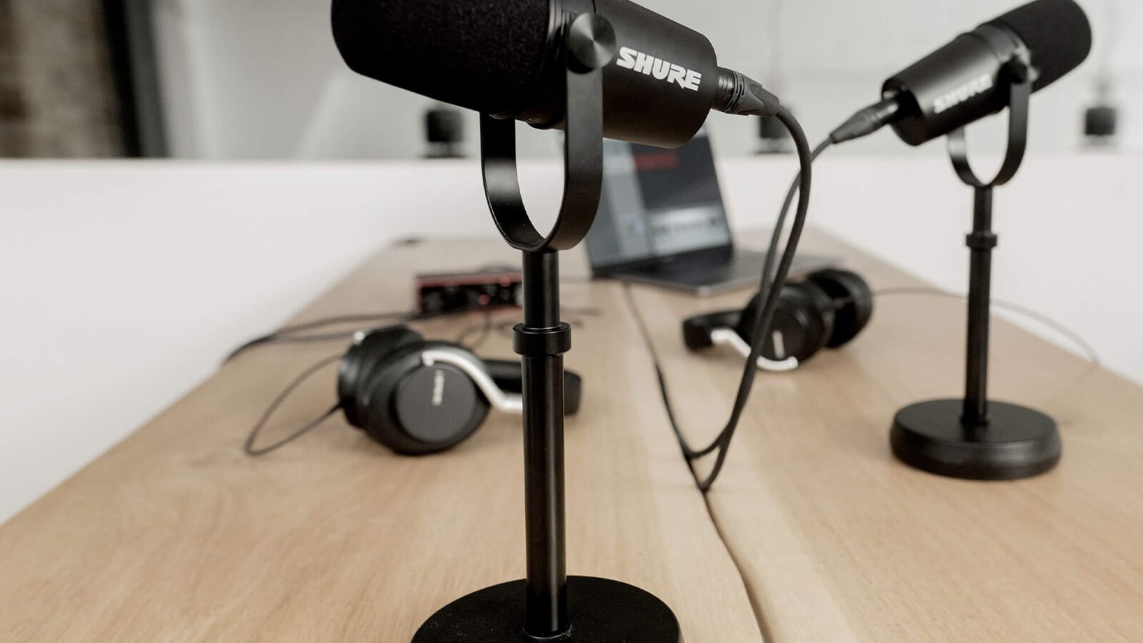 Dois microfones Shure MV7X pretos em pedestais de mesa, conectados por cabo XLR, sobre bancada de madeira com fones e interface de áudio