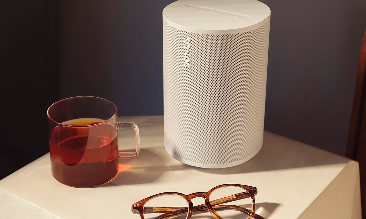 Caixa de som Sonos Era 100 branca sobre uma mesa lateral, ao lado de uma caneca de chá e um par de óculos