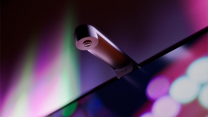 Câmera Philips Hue Play Screen Sync presa no topo de uma TV, com luzes coloridas roxas e verdes ao fundo, imagem oficial da Signify