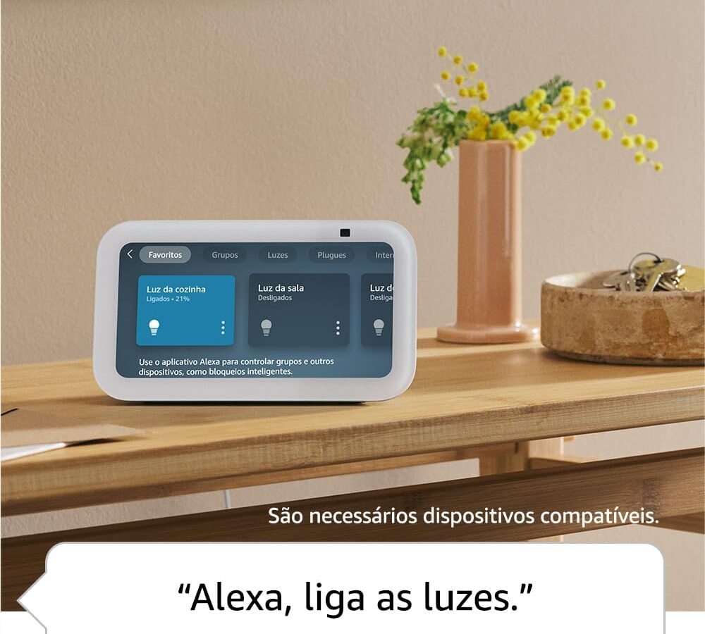 Echo Show 5 branco sobre um aparador de madeira ao lado de um vaso com flores amarelas, com a tela mostrando controles de luzes da casa e o balão de fala “Alexa, liga as luzes”