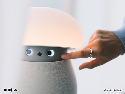 Robô doméstico OKA Mori, em formato de luminária com cúpula iluminada e dois olhos, sendo tocado por uma mão
