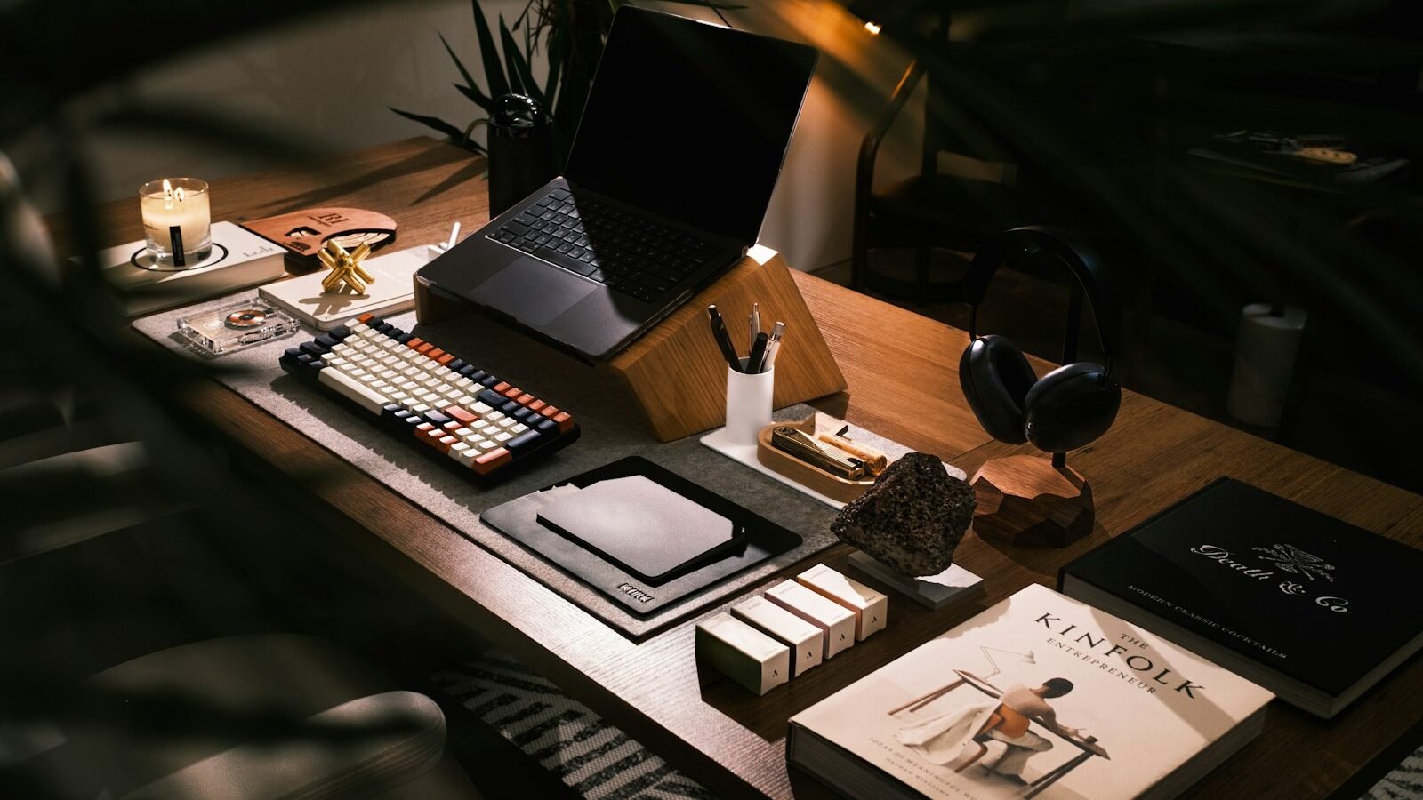 Mesa de home office com notebook em suporte de madeira, teclado, fone de ouvido e livros, sobre tapete em ambiente com plantas