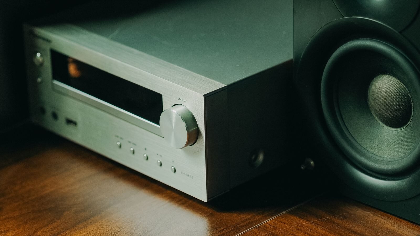 Amplificador estéreo prateado ao lado de uma caixa acústica passiva sobre piso de madeira