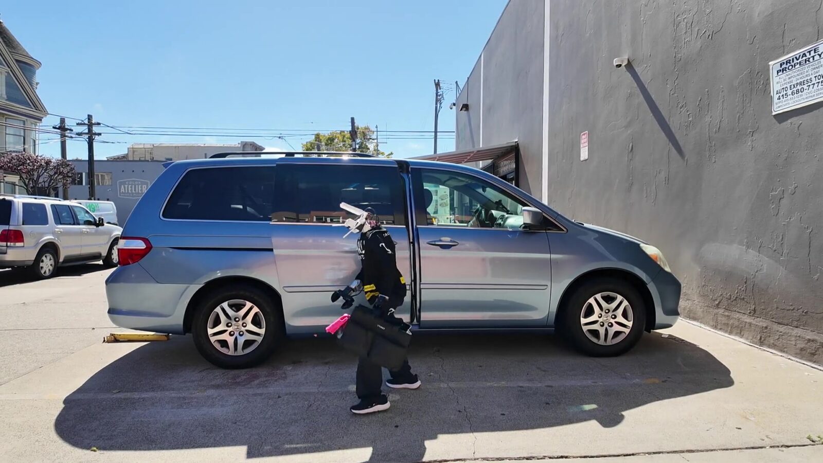 Robô humanoide da Tau Robotics, de roupa preta, carregando uma bolsa de material de limpeza ao lado de uma minivan prata estacionada em uma rua de San Francisco
