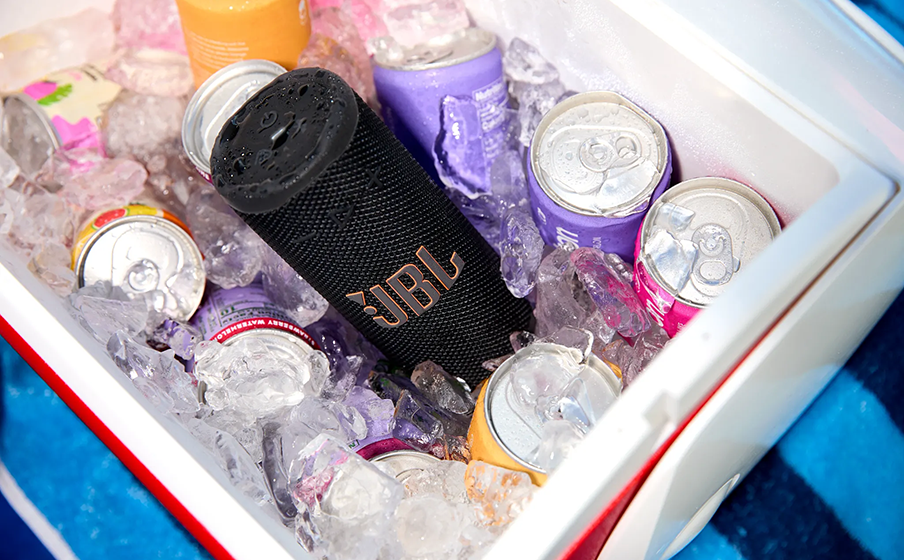 Caixa Bluetooth JBL Grip preta deitada entre latas de bebida e gelo dentro de um cooler