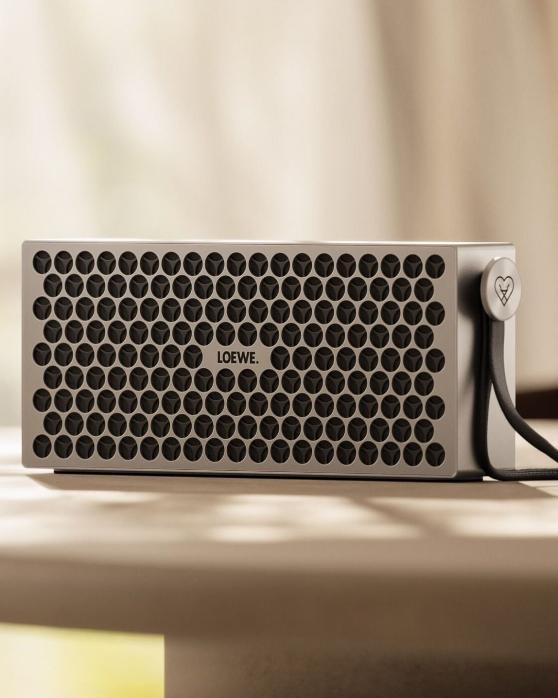 Caixa Bluetooth Loewe Neo em alumínio prateado com grade de furos circulares e alça de couro sobre uma mesa clara