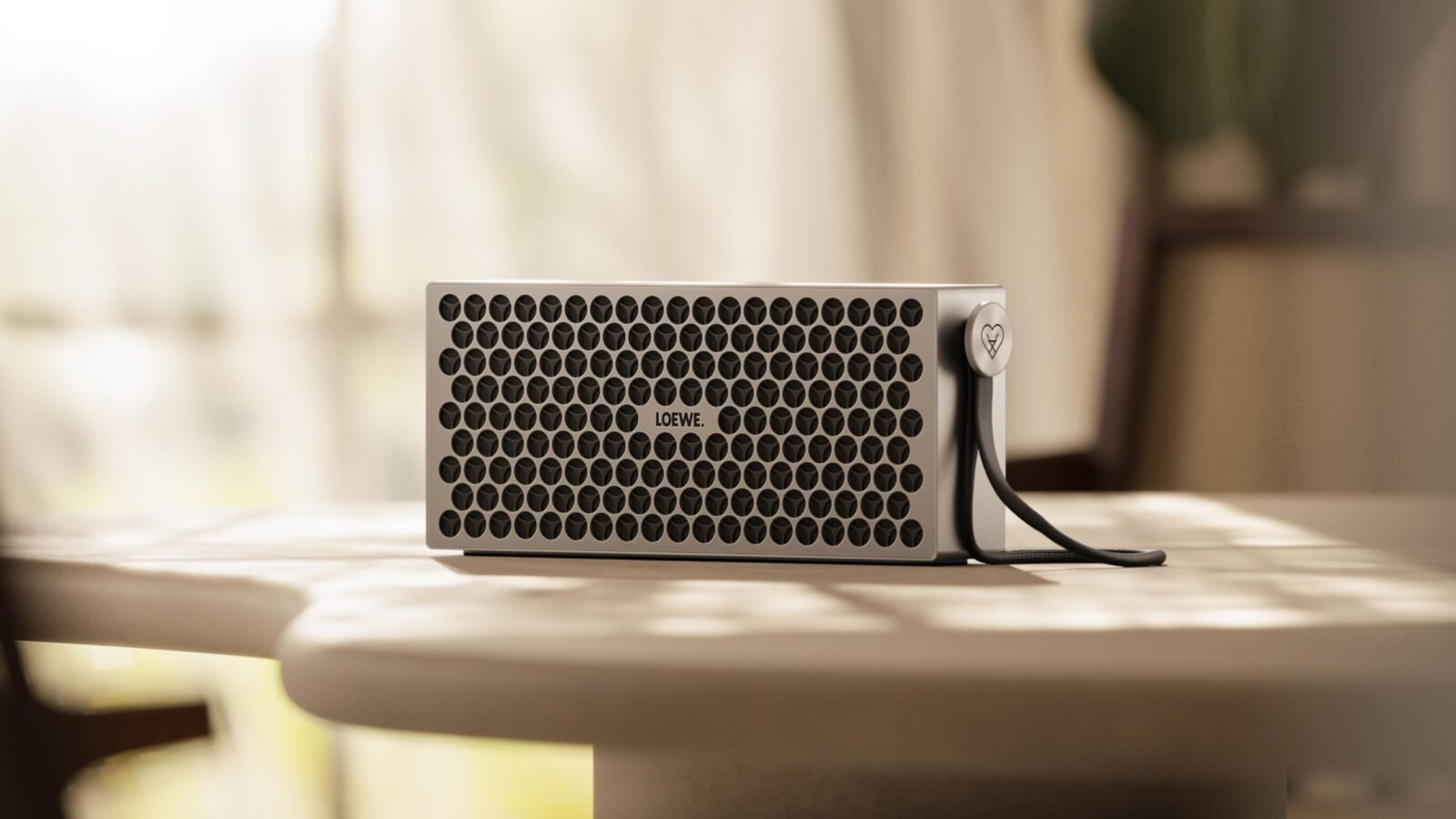 Caixa Bluetooth Loewe Neo em alumínio prateado com grade de furos circulares e alça de couro sobre uma mesa clara