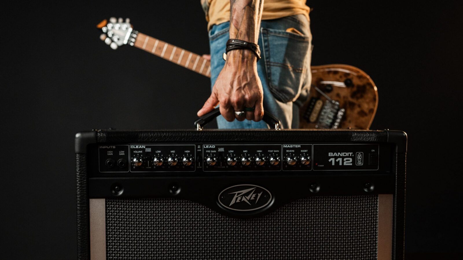 Músico carrega pela alça um amplificador combo Peavey Bandit 112 preto, com uma guitarra Peavey ao fundo