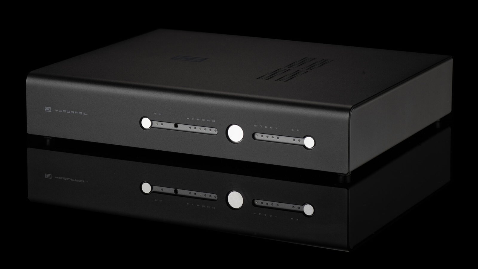 DAC Schiit Yggdrasil preto, com painel frontal de alumínio e botões prateados, sobre superfície reflexiva e fundo escuro